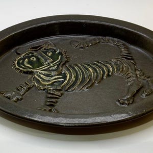 May include: A dark brown ceramic oval dish with a raised relief tiger design. The tiger has a green face and gold-colored stripes. The dish is a decorative item.