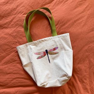May include: Off-white canvas tote bag with a dragonfly design. The dragonfly has pink, purple, and red wings and a black body. The bag has two side pockets and green handles.