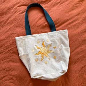 May include: White canvas tote bag with a sun and star design in yellow and orange. The tote has teal handles and two side pockets. The bag is sitting on an orange surface.