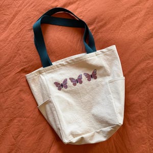 May include: Cream-colored tote bag with teal handles and three butterfly designs. The tote has two side pockets and is made of a sturdy canvas material. The butterflies are purple with a detailed wing pattern.
