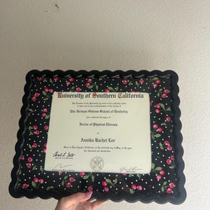 Puede incluir: Un diploma enmarcado de la Universidad del Sur de California, The Herman Ostrow School of Dentistry, para un Doctor en Fisioterapia. El marco es negro con un borde festoneado y un patrón de lunares blancos y cerezas rojas.