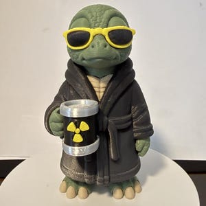 May include: A green reptilian figurine wearing bright yellow sunglasses and a dark gray bathrobe. The figure holds a black cylindrical container featuring a yellow radioactive hazard symbol with silver-colored top and bottom rims.