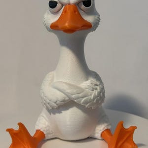 May include: A white goose figurine with a textured surface, featuring a bright orange beak and orange webbed feet. The goose has a grumpy facial expression with narrowed eyes and its wings are folded across its chest in a crossed-arm pose.
