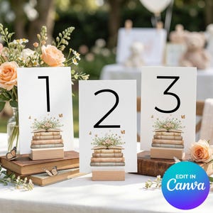 May include: White table number cards with black numerals 1, 2, and 3. Each card features a stack of books with floral and butterfly accents, set on a wooden base. The cards are arranged on a white tablecloth with flowers.