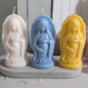 May include: Three pillar candles shaped like a pregnant figure with a halo, standing on a concrete base. The candles are colored white, light blue, and yellow, each featuring a wick at the top and a decorative rose pattern at the base.