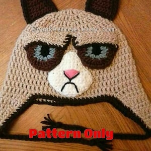 May include: A crocheted cat hat with brown ears, a white face, and a grumpy expression. The hat has a black and brown trim and a black string attached to the bottom. The text "Pattern Only" is written in red letters on the bottom of the image.