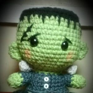 May include: Green crocheted Frankenstein monster plush toy with black hair, big eyes, and a stitched scar on its face. It is wearing a gray and black crocheted shirt with two white buttons.