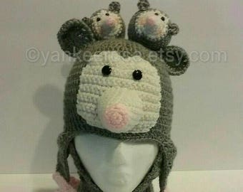 May include: Crochet pattern for a grey and white opossum hat with a pink nose and two smaller opossums on top. The hat is attached to a grey scarf with pink pom-poms. The pattern is for sale, not the finished product. "PATTERN ONLY" is written in blue text on the image.