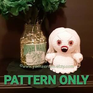 May include: A crochet pattern for a white ghost with red eyes and a red mouth. The ghost is sitting on a black surface with a green vase of flowers and a jar with a label that says "Irish Wake" in green letters.
