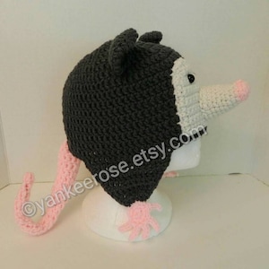 May include: A gray and white crocheted opossum hat with a pink tail. The hat has a white face with a black eye and a pink nose.