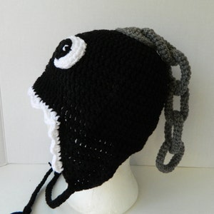 May include: Black crocheted hat with a white eye and a gray chain attached to the back. The hat has a white crocheted trim around the edge.