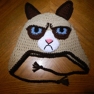 May include: A brown and white crocheted hat shaped like a cat with blue eyes and a pink nose. The hat has a brown crocheted brim and a brown crocheted tie.