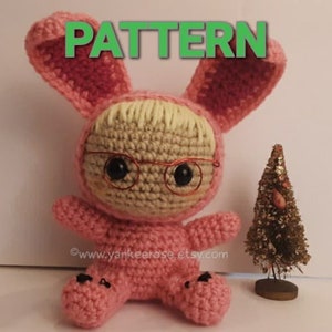 May include: Crocheted pink bunny rabbit wearing glasses. The bunny is sitting on a white surface. The text "PATTERN" is written in green letters above the bunny.