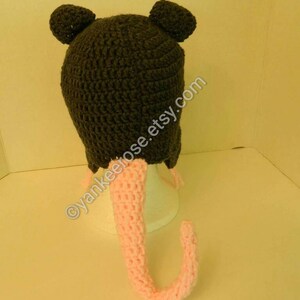 May include: A gray crocheted hat with pink crocheted ears and a long pink crocheted tail. The hat has a small, round pom-pom on each ear.