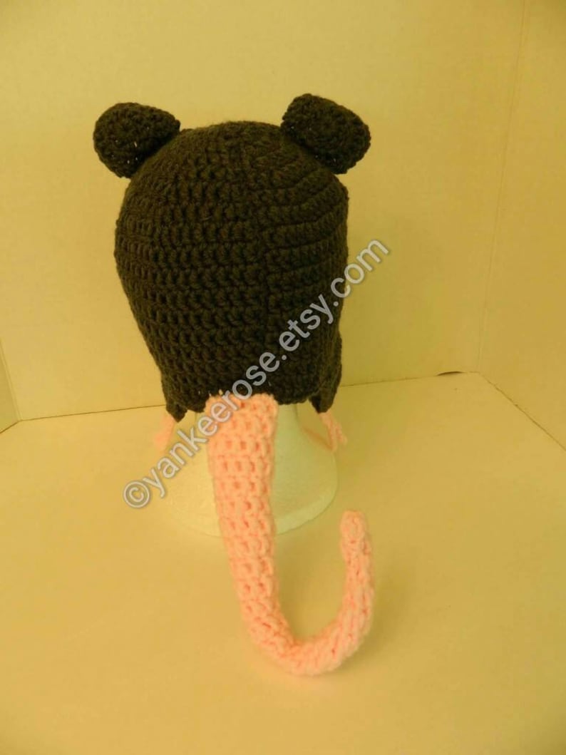 May include: A gray crocheted hat with pink crocheted ears and a long pink crocheted tail. The hat has a small, round pom-pom on each ear.