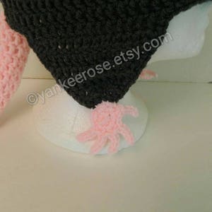 May include: A black crocheted hat with a pink crocheted tail. The hat is on a white mannequin head. The text "yankeerose.etsy.com" is on the hat.