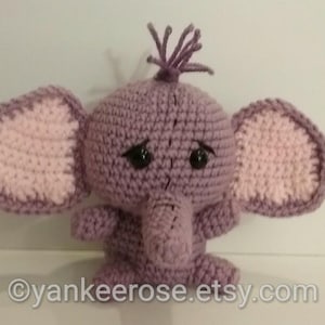 May include: A crocheted elephant plush toy with pink ears and a purple body. The elephant has black eyes and a sad expression.