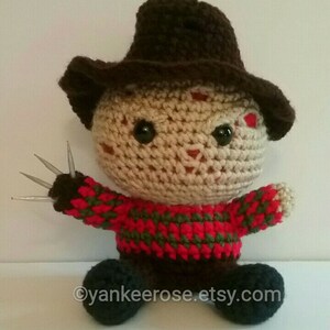 May include: A crocheted doll of a horror movie character, featuring a brown hat, a red and green striped sweater, and metal claws. The doll has a tan face with black eyes and a small red mark. The doll is sitting down.
