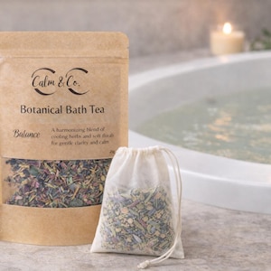 Puede incluir: Un paquete de té de baño botánico "Calm & Co.", con una ventana transparente que muestra la mezcla de hierbas. Una pequeña bolsa de muselina llena de hierbas está junto al paquete. El texto dice "Balance" y describe el té.