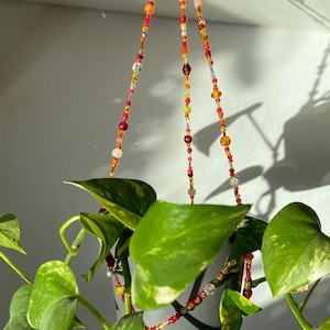 Könnte beinhalten: Ein hängender Blumentopf mit üppigen grünen Blättern und einem Perlenaufhänger. Die Perlen sind in Rot-, Orange-, Gelb- und Klartönen gehalten und bilden einen farbenfrohen und dekorativen Akzent. Der Blumentopf ist weiß.