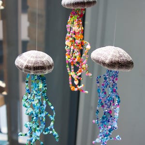Puede incluir: Tres decoraciones colgantes de medusas. Cada una tiene una parte superior redonda, texturizada y gris y coloridos tentáculos de cuentas en tonos de azul, verde, rosa, naranja y amarillo. Las medusas están suspendidas por finos hilos transparentes.