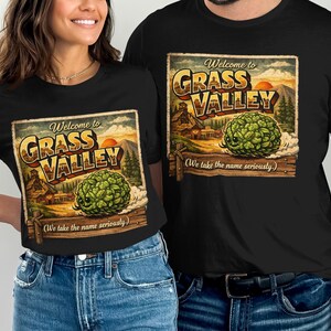 May include: Black t-shirts with a vintage-style graphic that reads "Welcome to Grass Valley." The design features a green, leafy plant, a sunset, and a mountain scene. The text "(We take the name seriously.)" is also included.