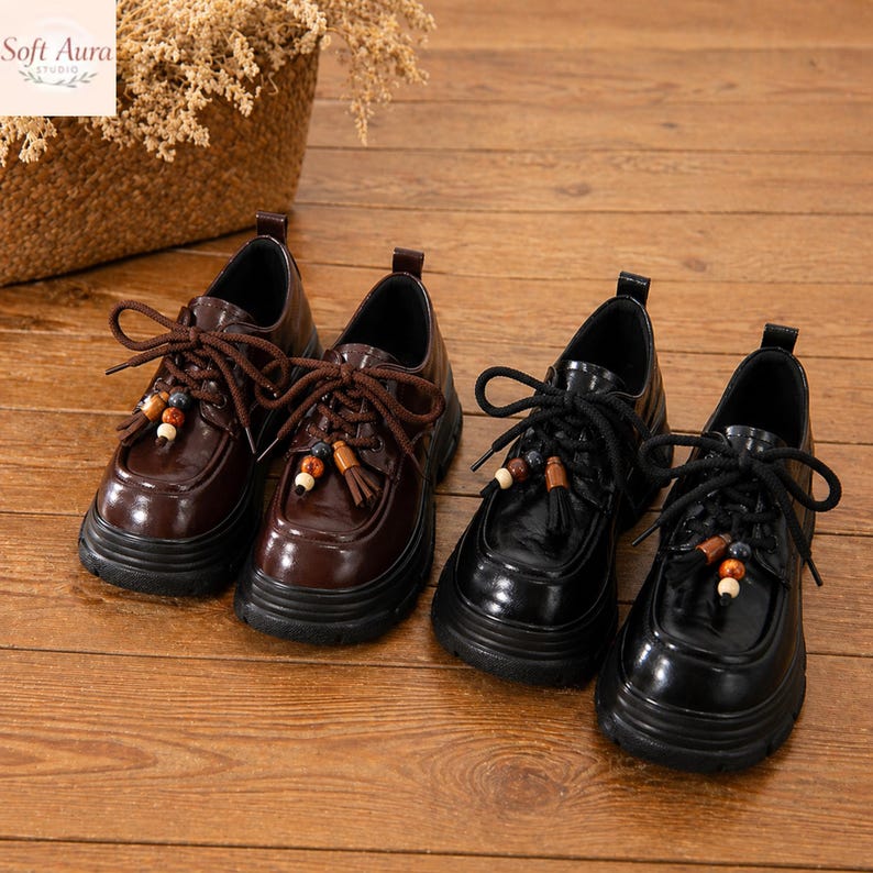 May include: Two pairs of oxfords, one in dark brown and the other in black, with thick soles. The brown shoes have brown laces and tassels with beads. The black shoes have black laces and tassels with beads. The shoes are on a wooden surface.