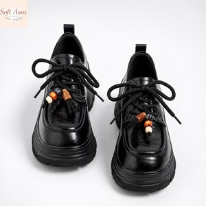 May include: Pair of black leather shoes with thick soles and decorative tassels. The shoes have black laces and are adorned with wooden beads. The shoes are a stylish and unique footwear option.