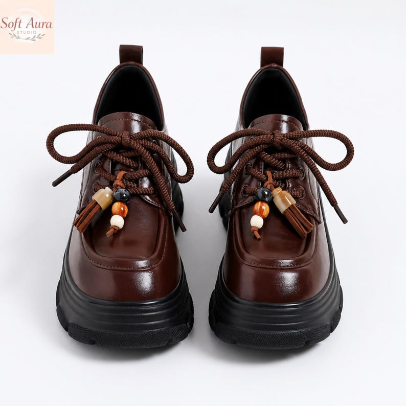 May include: A pair of brown leather shoes with thick black soles. The shoes have brown laces and decorative tassels with beads. The shoes are displayed on a white background. The brand name "Soft Aura" is in the top left corner.