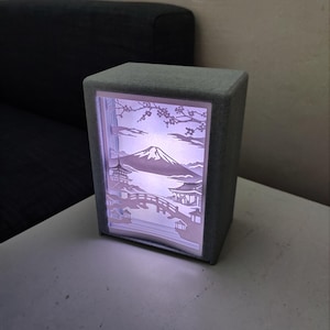 Pode incluir: Uma lâmpada retangular cinza com uma cena de paisagem japonesa iluminada no interior. A cena apresenta uma montanha, uma ponte e edifícios. A lâmpada está sobre uma superfície branca.