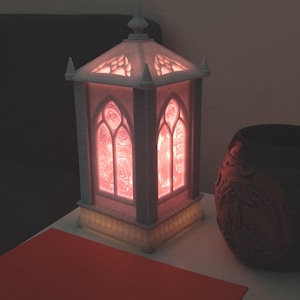 May include: A rectangular lantern with a light gray frame and a pointed roof, featuring translucent panels that glow with a warm pink light. The panels display intricate cut-out designs of an owl and architectural elements in a gothic style.
