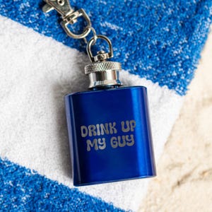 May include: A small, cobalt blue flask with a silver cap and attached keychain. The flask has the phrase "DRINK UP MY GUY" etched in a light color. The flask is resting on a blue and white striped towel.