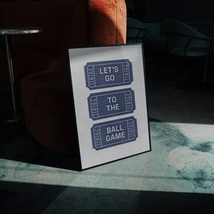 A framed vintage halftone print featuring three blue ticket stubs with the text "LET'S GO TO THE BALL GAME" in grey. The print is displayed on a marble floor, with a dark background and a glimpse of furniture.