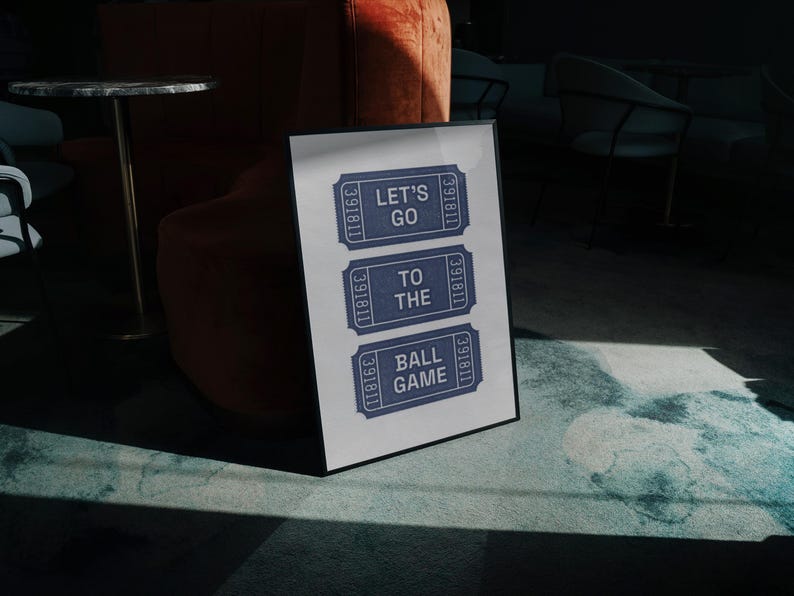 A framed vintage halftone print featuring three blue ticket stubs with the text "LET'S GO TO THE BALL GAME" in grey. The print is displayed on a marble floor, with a dark background and a glimpse of furniture.