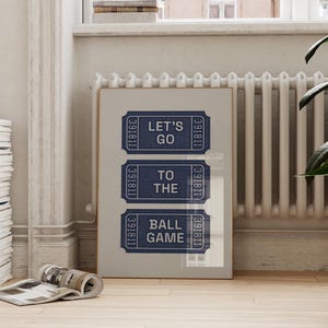 A framed print featuring three navy blue ticket stubs with the text "LET'S GO TO THE BALL GAME" in white. The print is displayed against a light gray background, with a wooden frame. The image is set in a room with a radiator and a potted plant.
