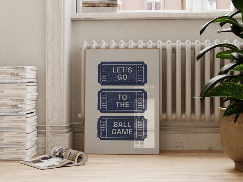 A framed print featuring three navy blue ticket stubs with the text "LET'S GO TO THE BALL GAME" in white. The print is displayed against a light gray background, with a wooden frame. The image is set in a room with a radiator and a potted plant.