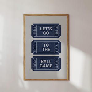 A framed print featuring three navy blue ticket stubs with white text. The top stub reads "LET'S GO", the middle reads "TO THE", and the bottom reads "BALL GAME". The frame is a light brown wooden oak.