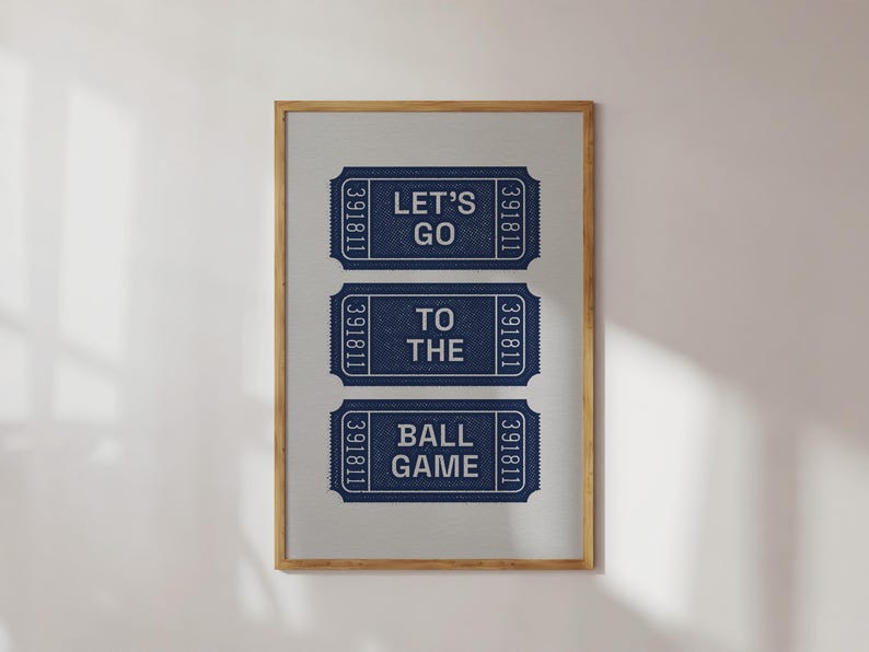A framed print featuring three navy blue ticket stubs with white text. The top stub reads "LET'S GO", the middle reads "TO THE", and the bottom reads "BALL GAME". The frame is a light brown wooden oak.