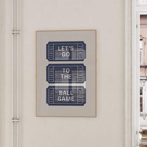 A framed print featuring three blue and white ticket stubs. The text on the tickets reads "LET'S GO TO THE BALL GAME." The print is displayed on a white wall near a window, with a glimpse of a building exterior.