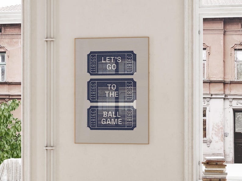 A framed print featuring three blue and white ticket stubs. The text on the tickets reads "LET'S GO TO THE BALL GAME." The print is displayed on a white wall near a window, with a glimpse of a building exterior.