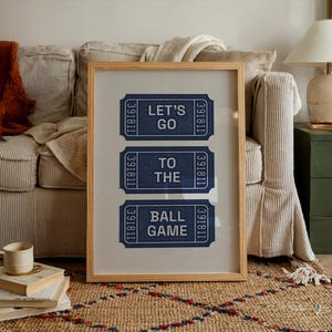 A framed print featuring three blue ticket stubs with white text that reads "LET'S GO TO THE BALL GAME." The tickets have a textured appearance and are set against a neutral background within a light wood frame. The print is displayed in a cozy living room setting.