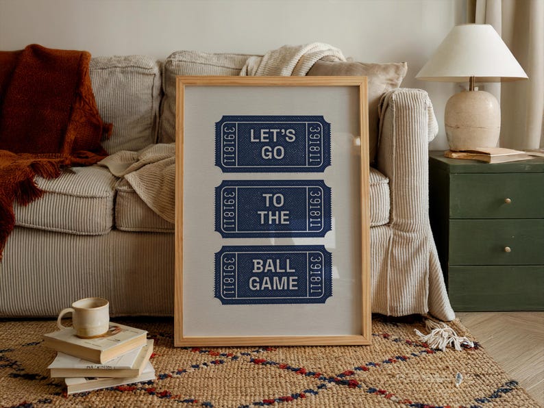 A framed print featuring three blue ticket stubs with white text that reads "LET'S GO TO THE BALL GAME." The tickets have a textured appearance and are set against a neutral background within a light wood frame. The print is displayed in a cozy living room setting.
