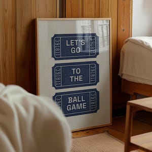 Framed vintage print featuring three navy blue ticket stubs with the text "LET'S GO TO THE BALL GAME" in white. The print is in a light wooden frame and leans against a wooden wall. The room has a bed and a small wooden table and bed sheets.