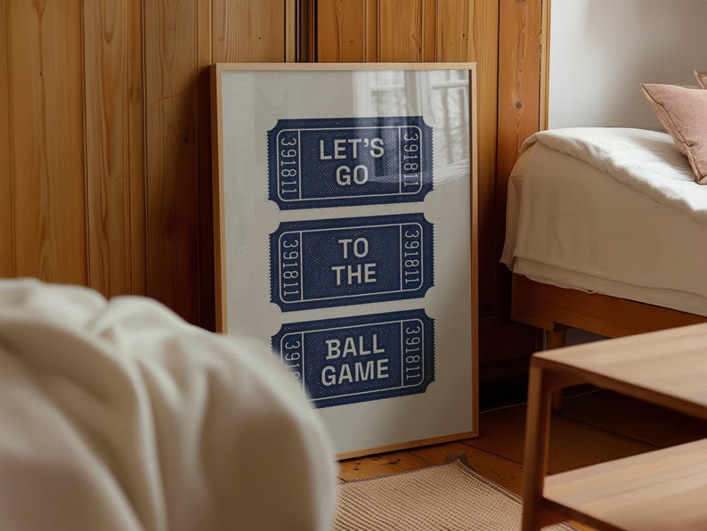 Framed vintage print featuring three navy blue ticket stubs with the text "LET'S GO TO THE BALL GAME" in white. The print is in a light wooden frame and leans against a wooden wall. The room has a bed and a small wooden table and bed sheets.