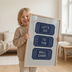 A child holding a framed vintage print featuring three navy blue ticket stubs with the text "LET'S GO TO THE BALL GAME". The tickets have a textured appearance and are set against a light gray background, framed in natural wood.