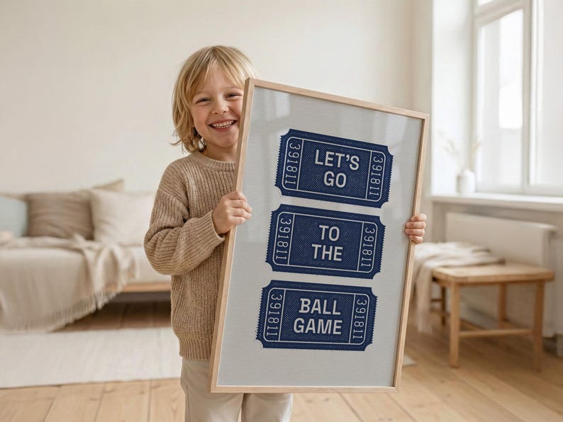 A child holding a framed vintage print featuring three navy blue ticket stubs with the text "LET'S GO TO THE BALL GAME". The tickets have a textured appearance and are set against a light gray background, framed in natural wood.