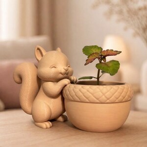 Op de afbeelding: Een beige eekhoornbeeldje lacht naast een kleine eikelvormige plantenbak. De plantenbak bevat een kleine plant met groene en rode bladeren. Het beeldje en de plantenbak staan op een houten oppervlak.