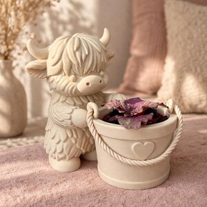 Peut inclure: Une figurine de vache des Highlands de couleur crème tenant un petit pot de fleurs en forme de seau avec une poignée en corde et un détail en forme de cœur. Le pot contient une plante violette. La figurine et le pot sont sur une surface rose.
