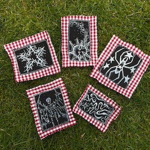 May include: Five black fabric patches with white illustrations and red and white gingham borders are arranged on green grass. The illustrations include a spiky ball, a spider, a skeleton, and abstract shapes. The patches are likely handmade.