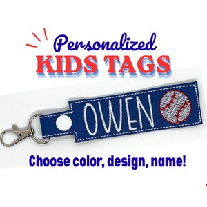 May include: A rectangular navy blue fabric tag featuring the embroidered white name OWEN and a baseball graphic. The tag includes a white snap button and a silver-toned metal lobster clasp. Text above reads Personalized KIDS TAGS and below reads Choose color, design, name!